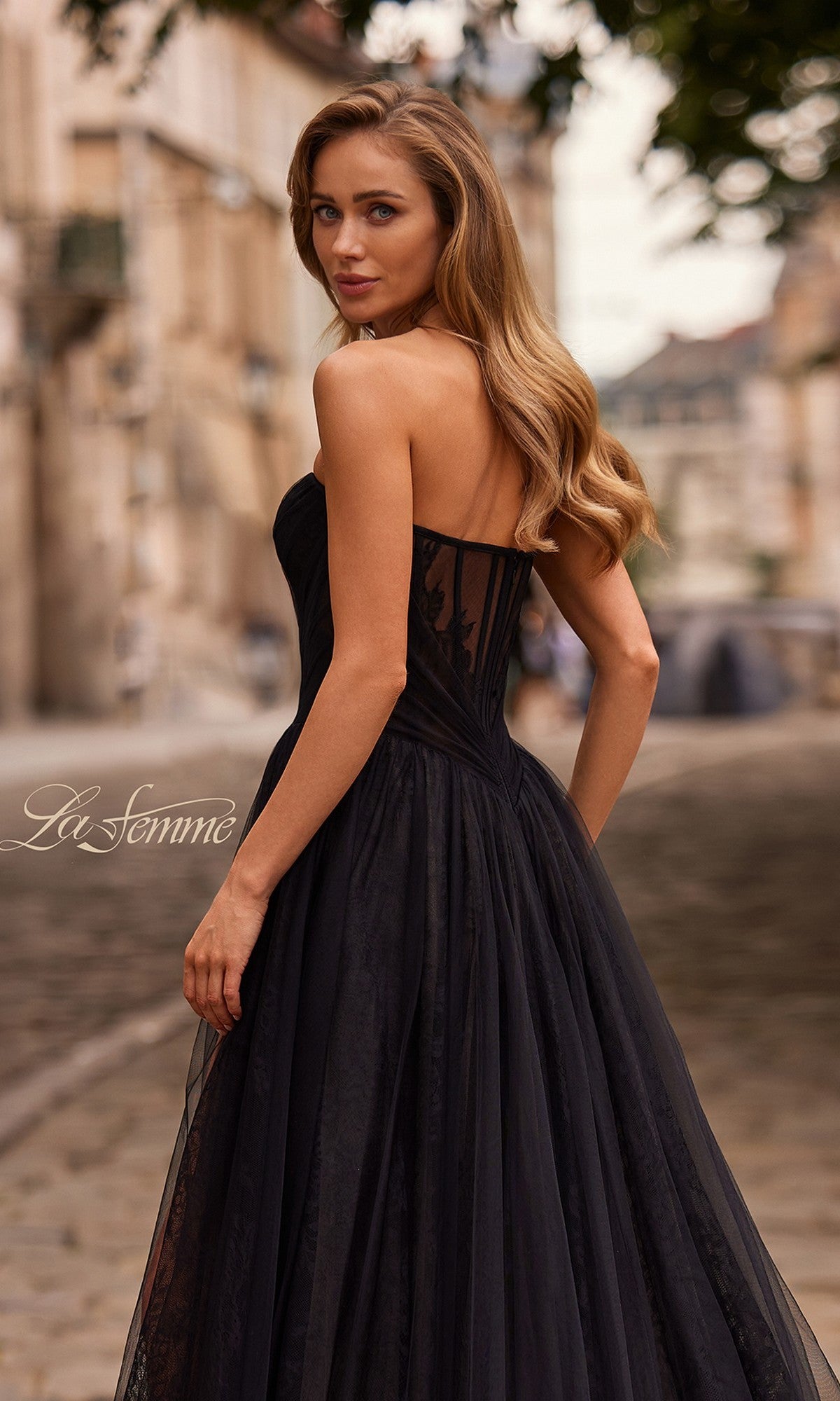 Woman with long wavy hair wears the La Femme 33307 long prom dress, standing outdoors on a cobblestone street and glancing over her shoulder.